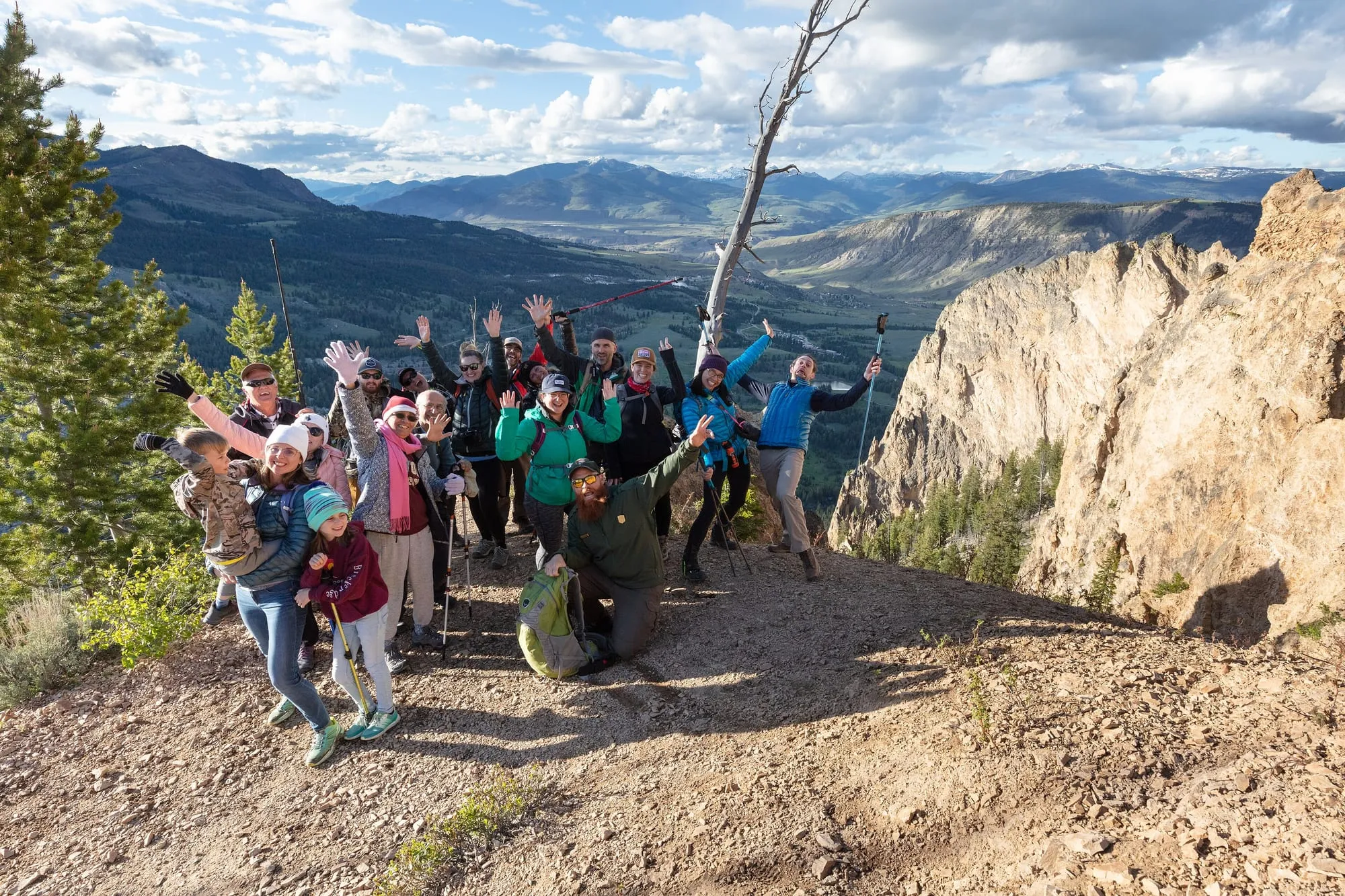 Group of smiling hikers waving with raised arms on a mountain trail in late-afternoon light